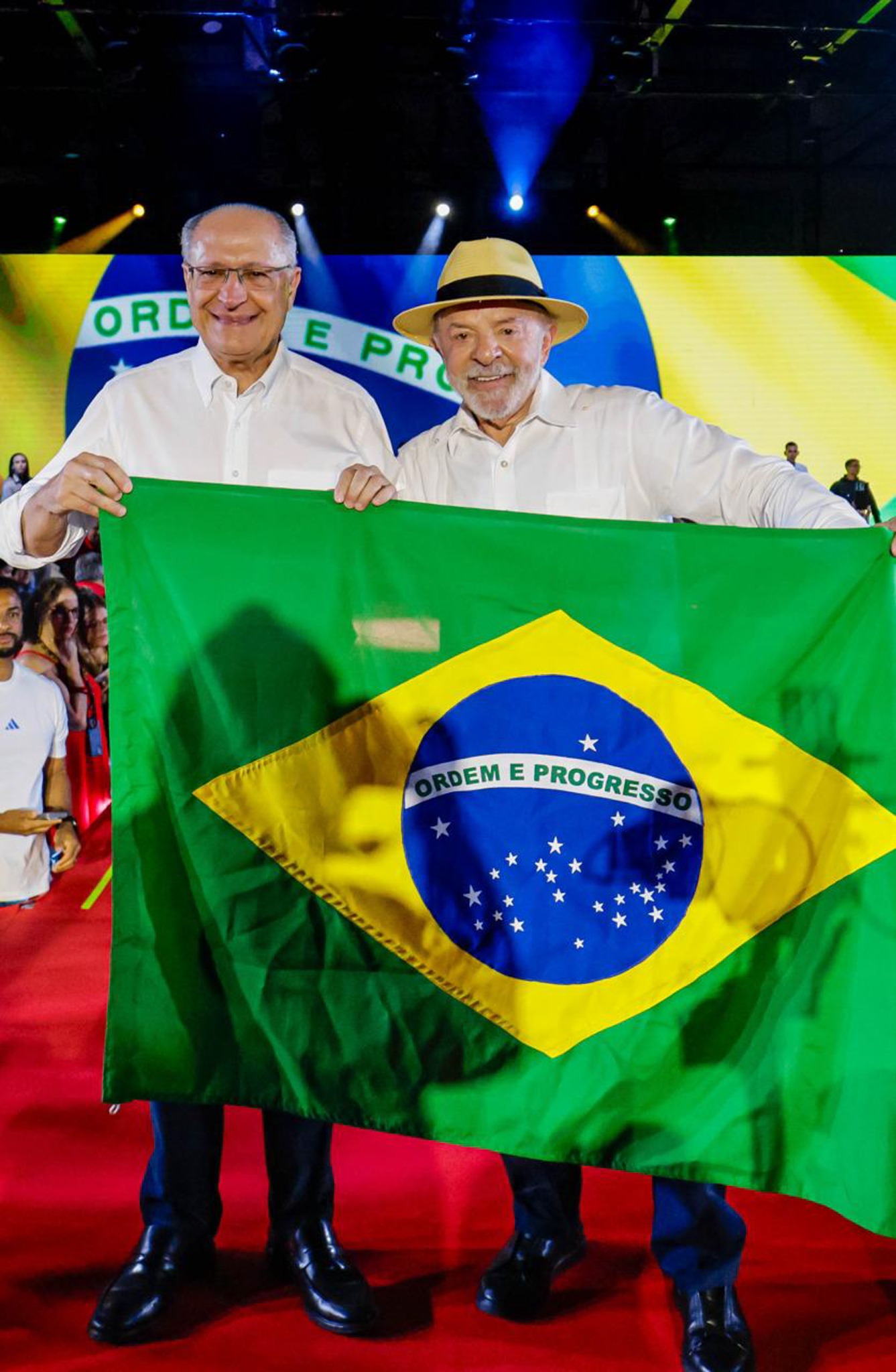 Geraldo Alckmin e Lula seguram uma bandeira do Brasil em um palco